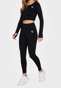 Czarny, długi rękaw, krótki top oraz legginsy z wysokim stanem, oba wykonane z elastycznego materiału, z logo na klatce piersiowej i udzie. Włożone z białymi sneakersami.