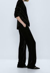 Veste en velours noir et pantalon ample assorti, présentant une texture lisse, une coupe ajustée et un éclat subtil. Des chaussures noires à bouts pointus complètent le look.