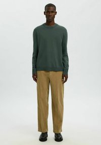 Hombre de pie mirando al frente, vestido con un suéter de manga larga verde oscuro, pantalones de pana beige y botas negras sobre un fondo simple.