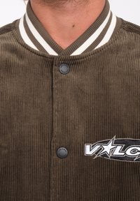 Bruine corduroy jas met een wit en grijs gestreepte ribkraag, drukknoopsluiting en geborduurd logo met witte en zwarte accenten.