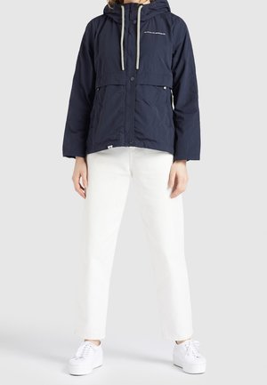 Frau trägt eine marineblaue Kapuzenjacke mit weißen Kordeln, weiße gerade geschnittene Hose und weiße Plateausneaker, steht vor einem schlichten Hintergrund.