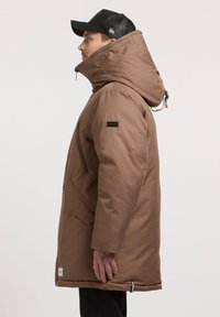 Braune, isolierte Jacke mit hohem Kragen und Kapuze. Verfügt über einen Frontreißverschluss und Seitentaschen. Schwarzes Logo-Label auf der Schulter.