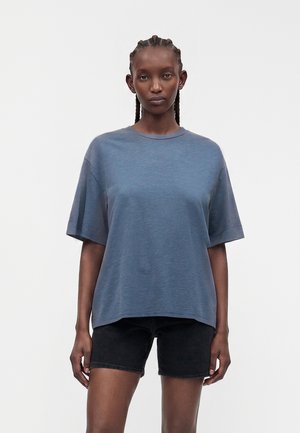 Femme aux cheveux tressés portant un t-shirt bleu ample et un short en jean noir, debout devant un fond clair uni.