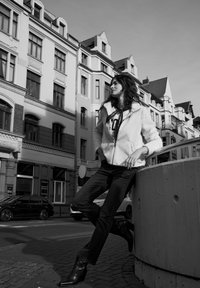 Modelka w jasnej, teksturowanej polarowej kurtce na zamek, nałożonej na wzorzystą koszulę, zestawiona z ciemnymi dżinsami i czarnymi botkami, opierająca się o ścianę.