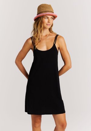 Mujer con vestido negro sin mangas con tirantes dobles y delgados y un sombrero marrón tejido con borde rosa, mirando hacia un lado.