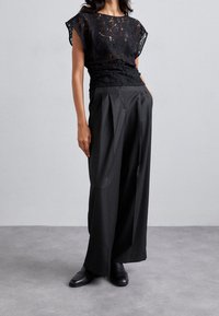 Femme portant un haut en dentelle noir à manches courtes, avec un pantalon plissé noir à jambes larges et des chaussures en cuir noires, se tenant devant un fond gris.