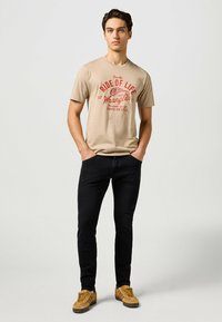 Vaatteena beige puuvillainen t-paita, jossa on punainen graafinen teksti, yhdistetty mustiin farkkuihin ja ruskeisiin tennareihin, joissa on teksturoitu muotoilu. Rennon istuvuuden omaava asu.