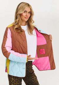 Žena ukazuje vnútornú kapsu prešívanej viacfarebnej bundy s ružovými, hnedými, modrými a žltými panelmi, oblečená v nohaviciach s leopardím vzorom.