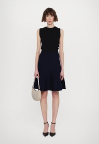 Haut sans manches noir, jupe évasée bleu marine, escarpins pointus noirs, portant un sac à main beige. Texture lisse et design minimal.