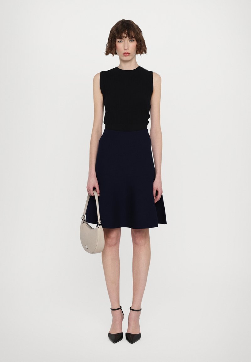 Haut sans manches noir, jupe évasée bleu marine, escarpins pointus noirs, portant un sac à main beige. Texture lisse et design minimal.