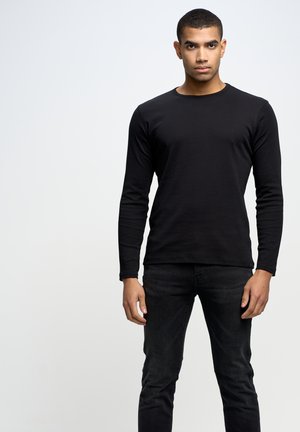 Camisa negra de manga larga, con cuello redondo y diseño ajustado. Combinada con jeans negros, de textura suave, con un detalle mínimo y sin logotipos visibles.