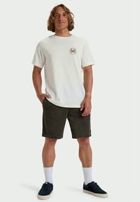 Jonge man staande met handen in zijn zakken, gekleed in een wit graphic t-shirt, donkere shorts, witte sokken en zwarte sportschoenen tegen een effen achtergrond.