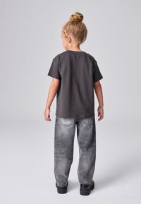 Malé dieťa so svetlými vlasmi zaviazanými do drdola, oblečené v čiernom tričku, voľných sivých džínsoch a čiernych topánkach, stojace chrbtom k divákovi na jednoduchom sivom pozadí.