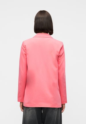 Personne aux cheveux courts et foncés portant un blazer rose vif et un pantalon gris foncé, vue de dos sur un fond clair uni.