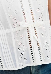 Weißes Baumwollbluse mit floralem Stickdesign, einer Knopfleiste und gebundenem Taillengürtel. Enthält Falten und schwarze Details entlang der Kanten.