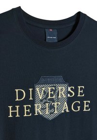 Granatowa koszulka z okrągłym dekoltem, z kremowym haftowanym wzorem, na którym widnieje napis "RÓŻNORODNE DZIEDZICTWO" oraz szczegóły w kształcie tarczy.