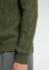 Tamsiai žalia megzta džemperis su tekstūruota kabelių raštu, raukiniais ant rankovių ir apykaklės. Medžiaga atrodo minkšta ir stora.