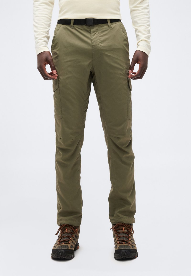 Άνδρας που φοράει χακί παντελόνι cargo, μαύρη ζώνη, μπεζ μακρυμάνικο πουκάμισο και καφέ ορειβατικά παπούτσια, στέκεται μπροστά σε μονόχρωμο φόντο.