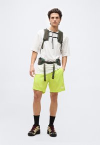 Νεαρός άνδρας που στέκεται, φοράει λευκό μπλουζάκι, σορτς νέον πράσινο, μαύρες κάλτσες, παπούτσια πεζοπορίας και ζώνη θώρακα και μέσης σε ελιά χρώμα.