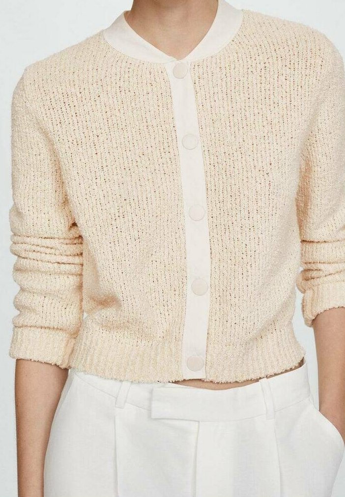 Jasno beżowy krótki kardigan z krótkim rękawem wykonany z teksturowanej dzianiny. Posiada okrągły biały kołnierz i dopasowane guziki z przodu. Krótki krój.