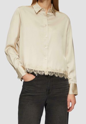 Woman wearing a cream satin button-up blouse with lace trim at hem, paired with high-waisted black jeans, one hand in pocket.