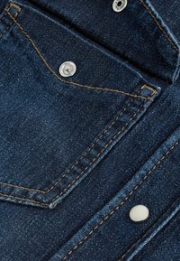 Țesătura de denim închis are o buzunar cusut pe piept cu un nasture metalic argintiu, fire contrastante de culoare maro și o textură netedă.