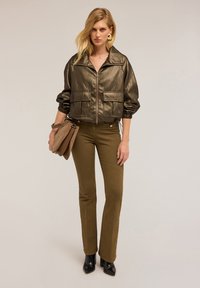 Giacca bomber verde oliva metallizzato con zip, due tasche frontali e polsini arricciati; abbinata a pantaloni verdi oliva aderenti e stivaletti neri con tacco.