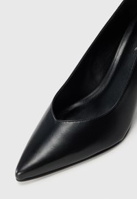 Chaussure à talon haut en cuir noir à bout pointu avec finition lisse, incliné pour montrer la partie supérieure et la zone interne du talon sur un fond clair.