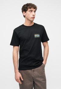 Άνδρας που φοράει μαύρο t-shirt Billabong με ένα μικρό πολύχρωμο τοπίο ως λογότυπο στο στήθος, συνδυασμένο με καφέ παντελόνι, στέκεται μπροστά σε λευκό φόντο.