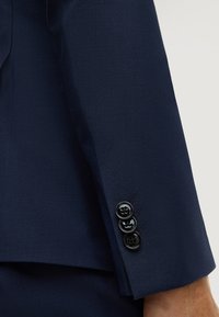 Manica di un abito blu navy con tre bottoni neri cuciti molto vicini e cuciture visibili dei bottoni sul braccio di una persona su uno sfondo chiaro.