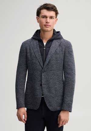 Mann trägt einen strukturierten grauen Blazer über einer schwarzen Kapuzenjacke mit Reißverschluss und dunkler Hose, steht vor einem schlichten hellen Hintergrund.