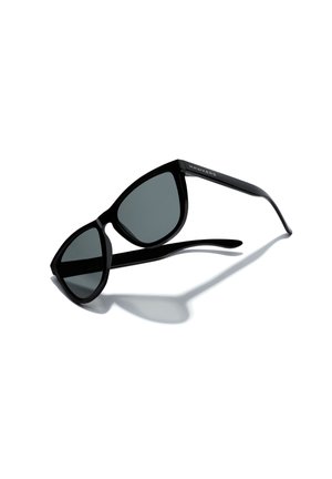 Black sunglasses with gray lenses, angular cat-eye shape, glossy plastic frame, and understated logo on the temple. Shadow cast on white background.