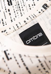 Krémové tričko s černým textovým vzorem, obsahující černou nášivku s logem "ombre" poblíž výstřihu. Měkk�á tkanina s hladkou texturou.