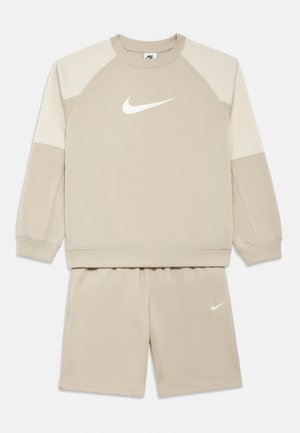 Bézs sportos szett hosszú ujjú, raglán ujjú, kerek nyakú pulóverrel és hozzáillő rövidnadrággal. Mindkét darabon fehér Nike logó található.