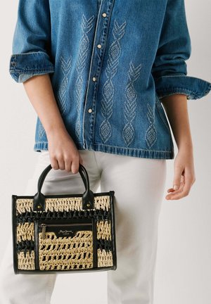 Persona sosteniendo un bolso tejido de paja y cuero negro, vistiendo una chaqueta vaquera azul bordada y pantalones blancos.