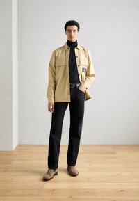 Beige Hemd mit Knöpfen über einem schwarzen Rollkragenpullover, schwarze Hose und braune Stiefeletten, mit einem Logo-Patch auf der linken Seite des Hemdes.