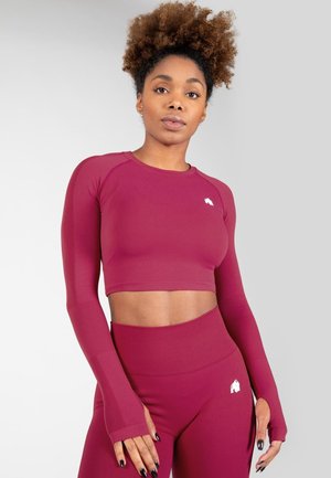 Burgunderfarbenes sportliches Crop-Top mit strukturierten Ärmeldetails und enganliegendem Design. Es verfügt über ein kleines weißes Logo auf der Brust und wird mit passenden Leggings kombiniert.
