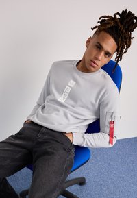 Felpa grigia con un patch logo, maniche lunghe e una tasca con zip sul braccio sinistro. Jeans neri e una sedia da ufficio blu visibili.