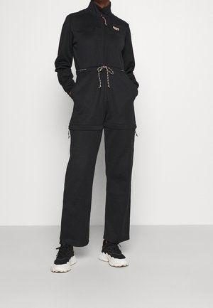 Ensemble de survêtement noir à manches longues, avec fermeture éclair à l'avant, cordon de serrage ajustable à la taille et deux poches zippées. Associé à des baskets noires et blanches épaisses.