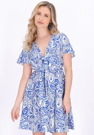 Femeie cu păr lung și blond poartă o rochie cu model floral albastru și alb, cu mâneci scurte, decolteu în V și cordon în față, stând în fața unui fundal alb simplu.