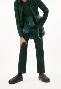 Persona che indossa pantaloni verde scuro, camicie sovrapposte verdi e blu e scarpe slip-on nere trapuntate, in piedi con una mano appoggiata su un ginocchio.