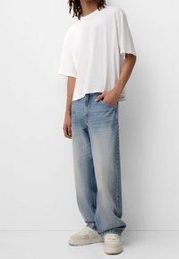 Vit kortärmad t-shirt i croppad modell, i kombination med lösa, ljusblå jeans och vita sneakers på en grå bakgrund.