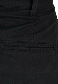 Gros plan sur un tissu noir montrant une poche fendue horizontale avec des coutures visibles près d'une boucle de ceinture sur un vêtement.