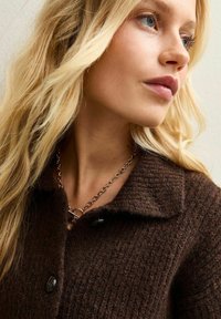 Donna bionda con occhi azzurri chiari, che indossa un cardigan di maglia marrone spesso e una collana d'oro, mentre guarda di lato su uno sfondo neutro.