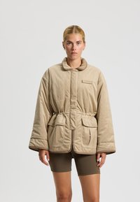 Chaqueta de pana beige con cuello alto, cintura ajustada y dos bolsillos frontales. Textura suave con detalles de ribete en contraste.