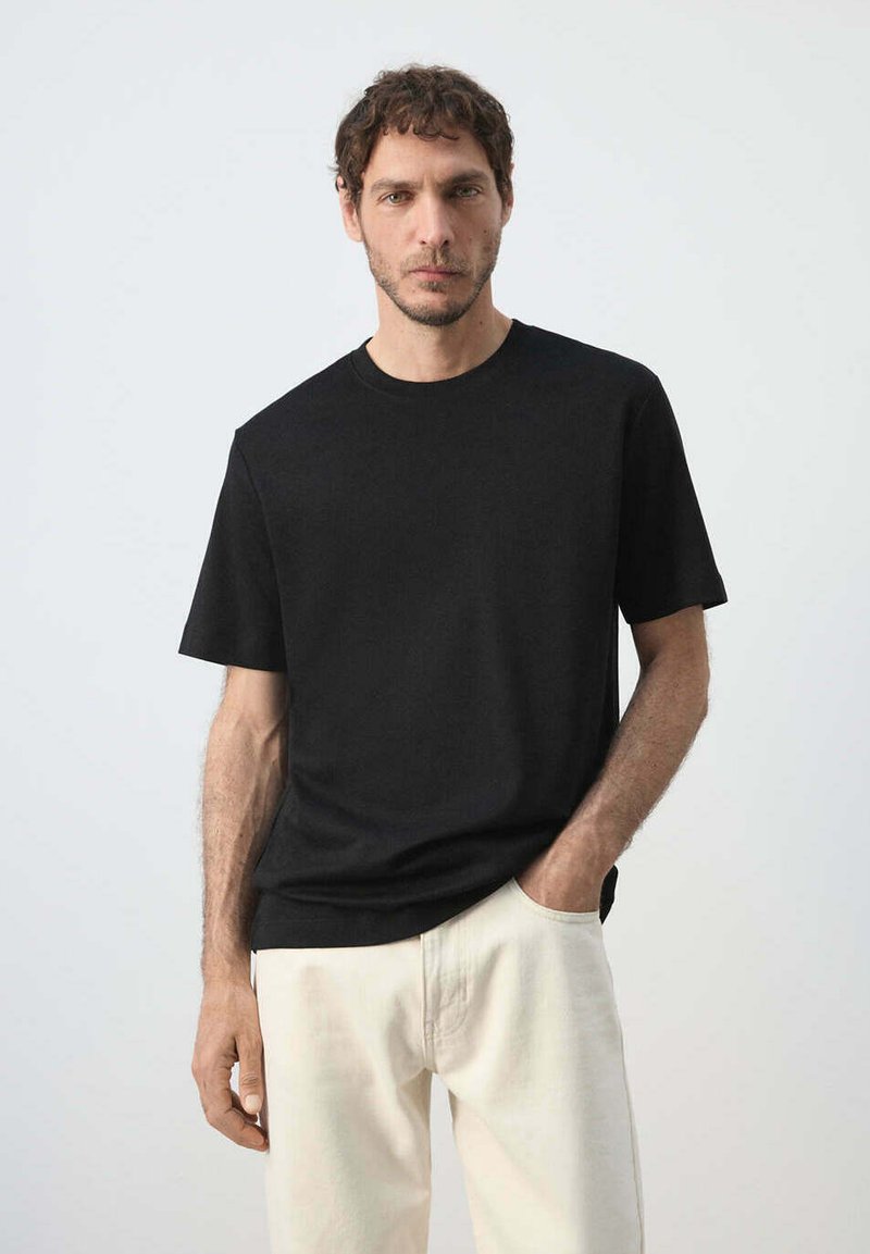 Homme aux cheveux courts et bouclés portant une chemise noire à manches courtes et un pantalon clair, debout avec une main dans la poche.