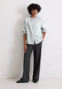 Jasnoniebieska koszula zapinana na guziki z długim rękawem i marszczonym dekoltem, zestawiona z szerokimi ciemnoszarymi spodniami, stoi na jasnym tle.