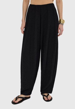 Žena nosí volné černé kalhoty, černé žabky a na pravém zápěstí zlato-stříbrné náramky, stojí na jednoduchém pozadí.