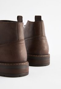 Hnedé kožené členkové čižmy s hladkou textúrou, okrúhlym špičkou a pútkom na ťahanie. Dizajn dopĺňa kontrastné prešívanie a drevený podpätok.