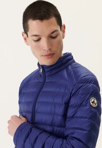Chaqueta acolchada azul con patrón de costura horizontal, cuello alto, cremallera frontal y parche de logo redondo en la manga izquierda. Textura suave y brillante.